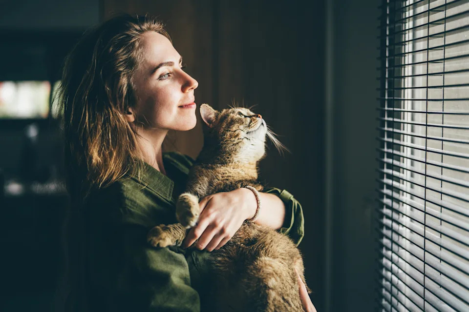 A woman smiles while holding a cat, both gazing out a window with blinds partially open