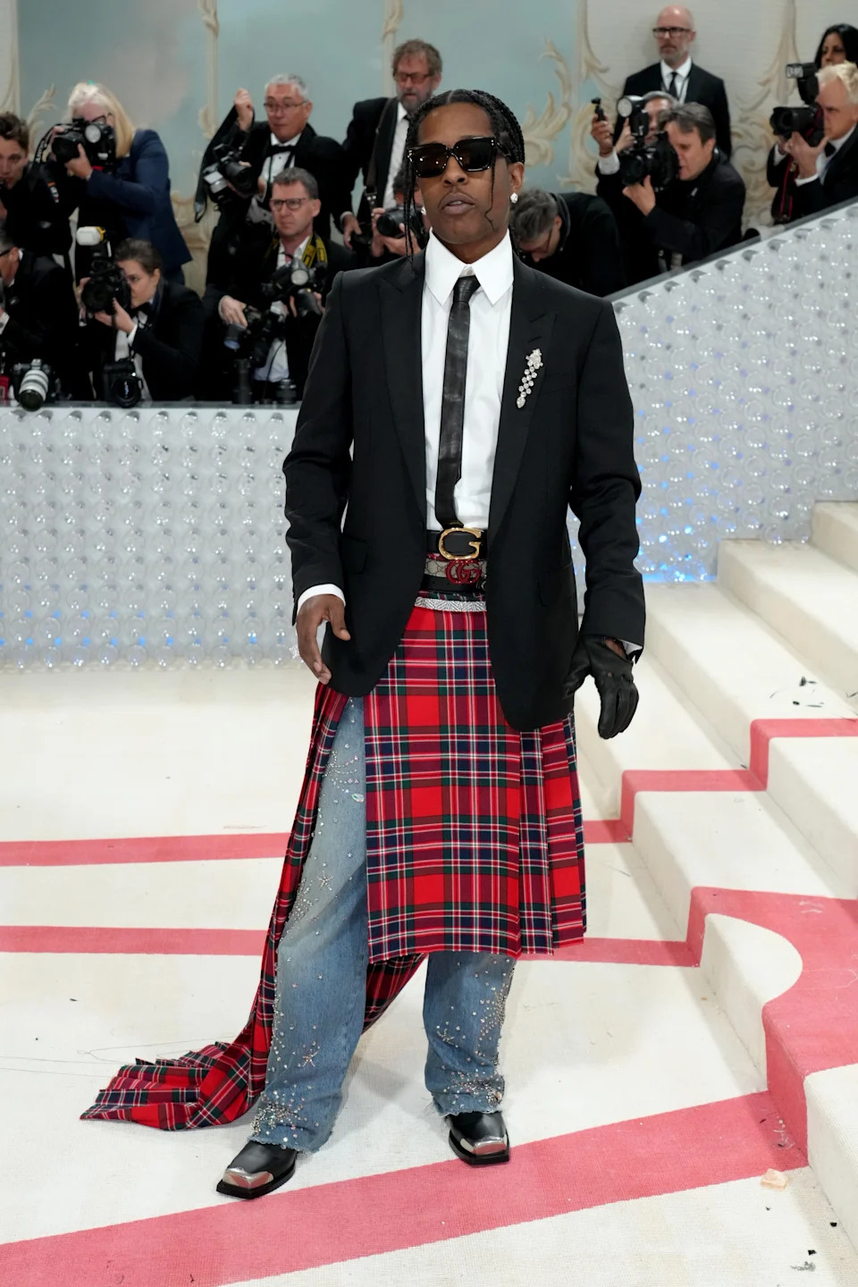 A person on a red carpet wears a black blazer, white shirt, tartan skirt, and jeans with embellishments, surrounded by photographers