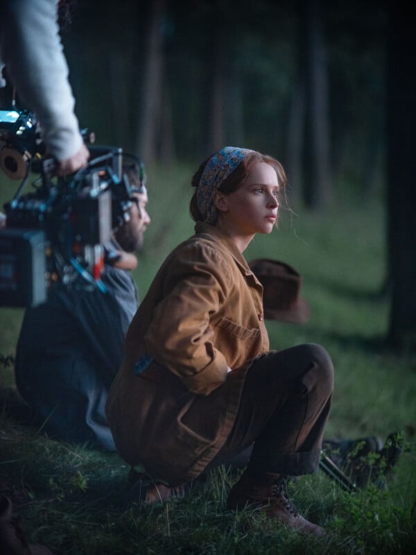 A woman wearing a brown jacket and headscarf sits on grass in a forest, looking thoughtful. A camera operator is next to her, suggesting a film is being shot.
