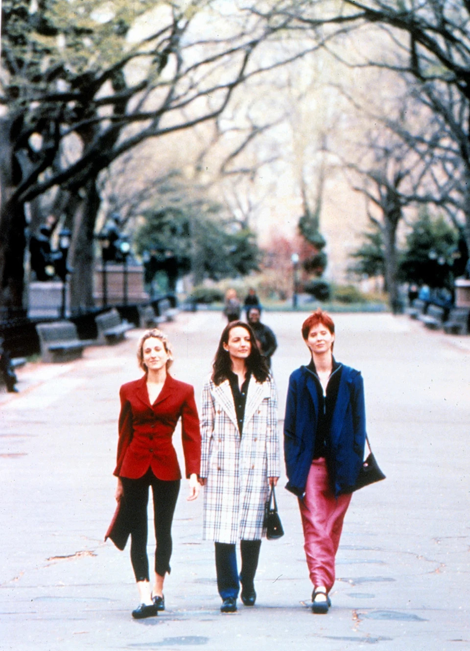 Three women are walking confidently in a park. One wears a red jacket, another a checkered coat, and the third a blue jacket with pink pants