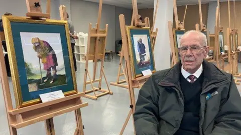 BBC Dick Scott, with balding grey hair and a moustache, wearing glasses, a green coat, navy jumper, white shirt and tie, sits in a chair next to a series of watercolour paintings on a wooden easels. 