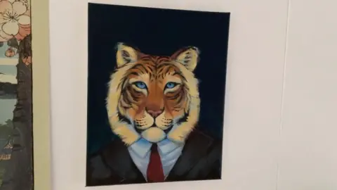 Aisha Belarbi A picture hanging on a white wall. It shows a tiger's head, with striped fur and piercing blue eyes, sitting on top of a dark suit, white shirt and red tie.