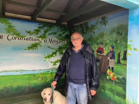 A man wearing a dark raincoat, standing with a golden labrador dog in a bus shelter with artwork on the walls behind him
