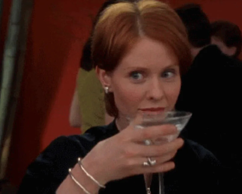 A person with a short hairstyle holds a martini glass, looking off to the side with a playful expression