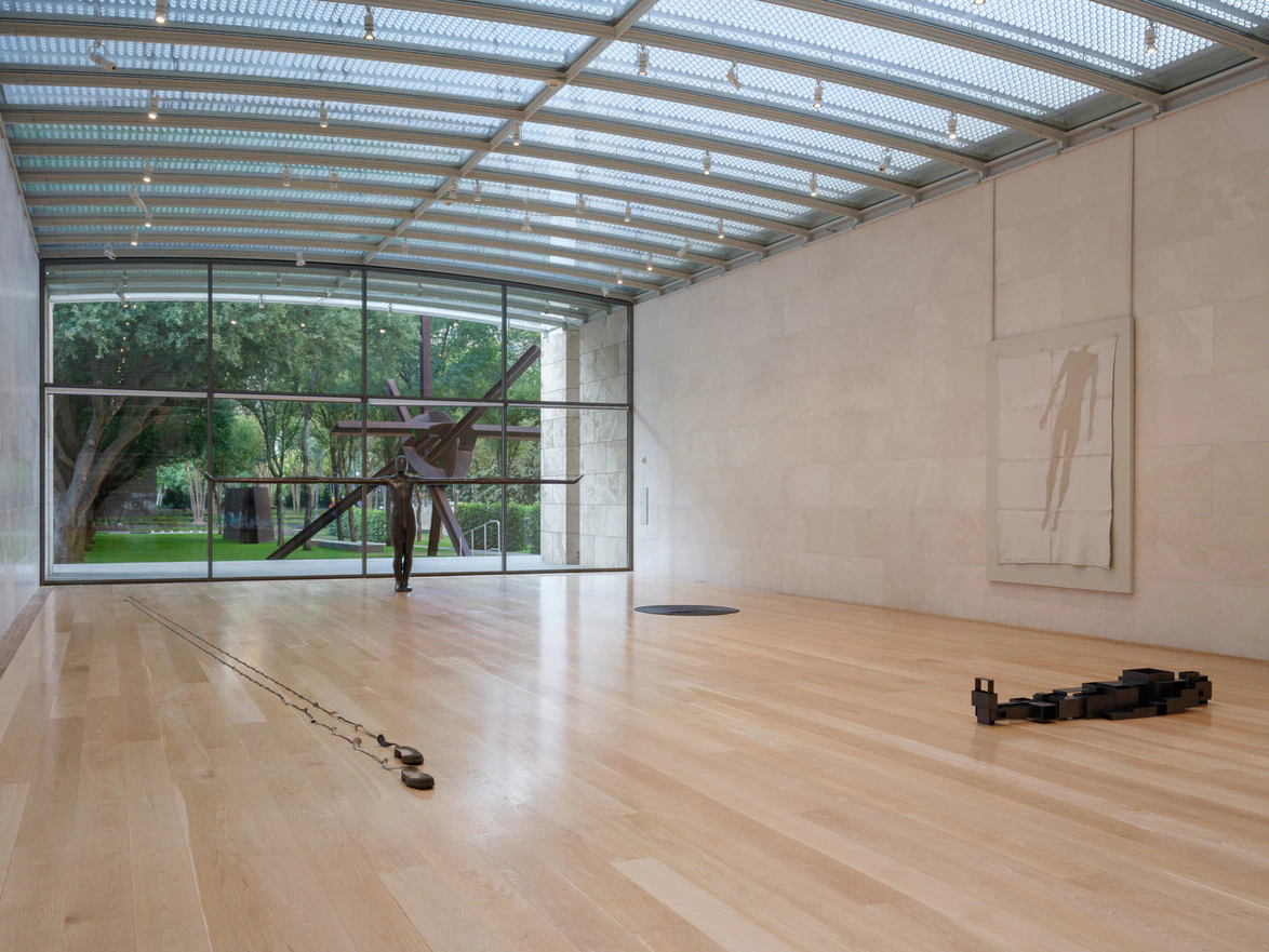 Modern art gallery with wooden floors, large sculpture near window, abstract painting on wall, and open view of garden outside.