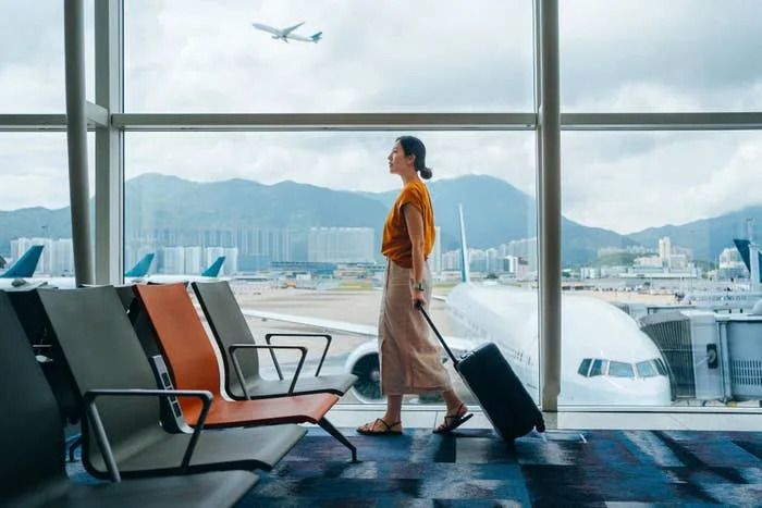 Woman walking with luggage in an airport terminal, large windows show airplane taking off and cityscape in the background