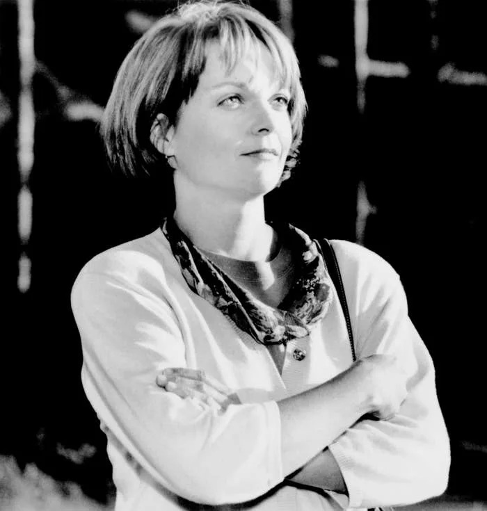 Person with short hair crosses arms, wearing a scarf and jacket, gazing optimistically. Appears thoughtful, scene suggests contemplation