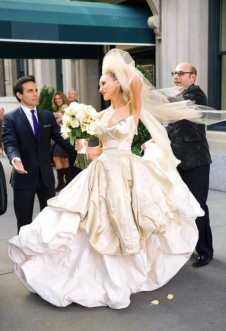 A woman in an elaborate gown holds a bouquet, adjusting her long veil as two men assist her outside a building, capturing a glamorous, bridal moment