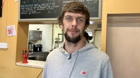 A man with brown hair and a bed in a grey hoodie. He is standing in front of a service hatch