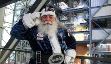 A man with a big white beard wearing a blue Santa costume with Artemis and NASA patches points to his hat. The Artemis II SLS and Orion spacecraft are In the background.