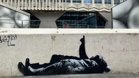 The black and white street art depicts two children lying on their backs and pointing upwards. The child nearest the camera is wearing a wooly hat with hands tucked into their jacket pockets. Behind them a larger person is pointing upwards in a bobbly hat (possibly a Santa type hat) also wearing boots.