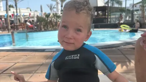 Contributed Edward stands in a blue, black and grey wetsuit next to a swimming pool with two people in it. He has dark blonde wavy hair.