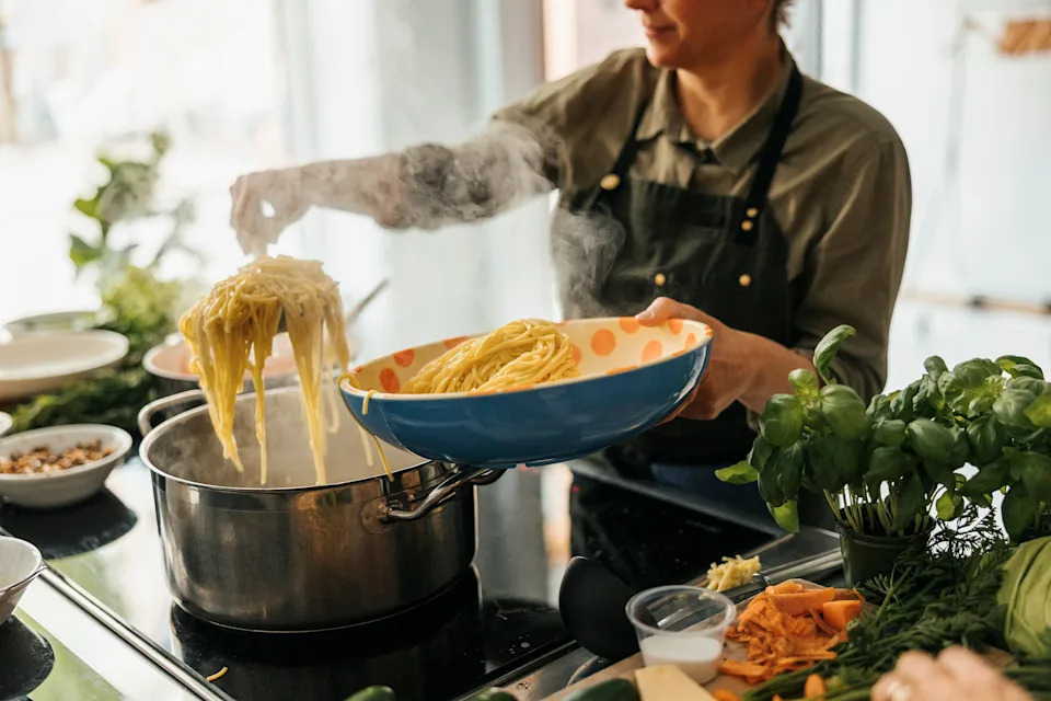 Person cooking pasta over a stove, holding pasta with tongs above a pot, surrounded by fresh ingredients on the counter