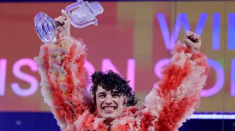 Reuters Swiss singer Nemo holds up the 2024 Eurovision trophy on stage wearing a fluffy, pink ruffled jacket.
