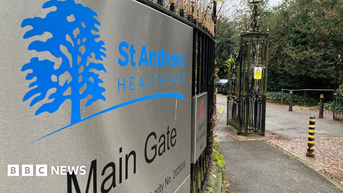 Main gate of St Andrew's Healthcare. There is a metal sign on black railings, with blue logo shaped like a tree. To the right is an iron fence and some black and yellow striped bollards.