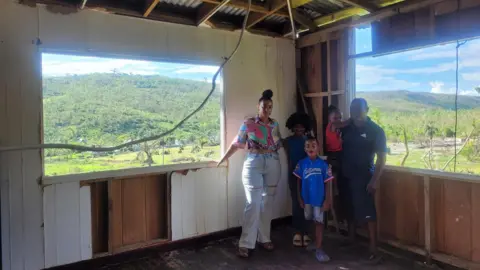 Handout A picture of Sabrina and her husband and three young children in their home. The windows are gone and a wire is hanging from the ceiling. The wooden boards of the wall have come off.