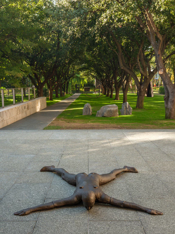 Bronze sculpture of a human figure lying face down on a stone path lined with trees and green grass in a park setting.