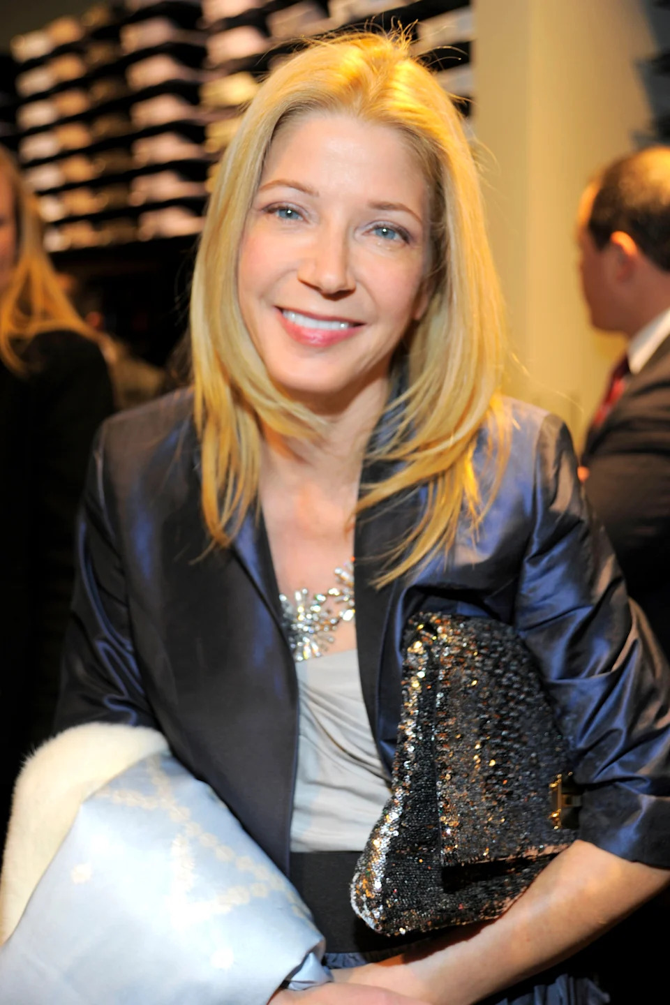 Person smiling, wearing a sleek jacket and holding a textured clutch. Background shows an indoor event with people and shelves
