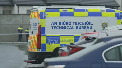 RTÉ A white van with yellow and blue chequered paint has the words 'AN BIÚRÓ TEICNIÚIL. TECHNICAL BUREAU written on its side. Beyond the van is white and blue striped police tape and two men wearing black and luminous yellow jackets. 
