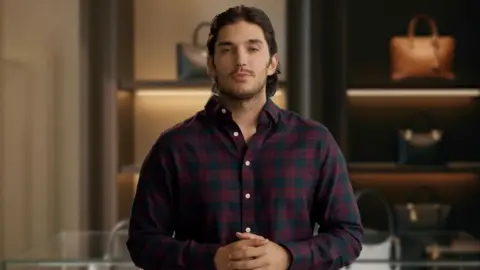 MirrorMe AI An AI-generated image that shows a man standing inside a handbag shop, wearing a long-sleeved checked shirt. His hands are clasped in front of him and he appears to be speaking. His shoulder-length dark hair is brushed back over his head and he has dark stubble on his face. Behind him are handbags arranged on shelves. They each have their own shelf and are individually lit.