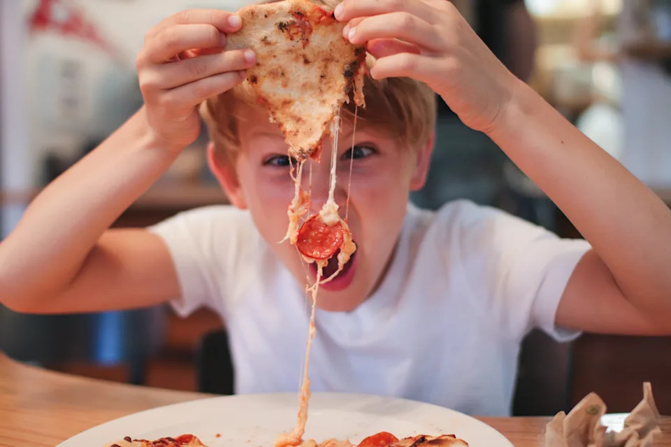 Boy messily eats cheesy slice of pepperoni pizza, pulling it upwards