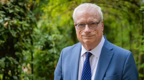 PA Lord Chris Smith is wearing a blue suite and tie and is standing in green leafy gardens.