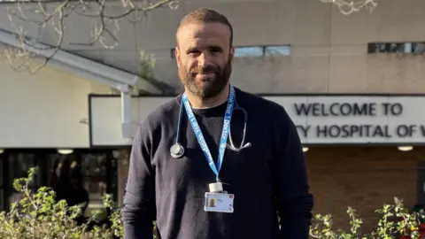 Cardiff and Vale University Health Board Jamie Roberts stood out the UHW Hospital in Cardiff. He is smiling wearing a navy blue jumper with his lanyard and stethoscope around his neck.