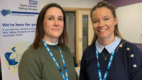 BBC/Seb Cheer Two women stand looking at the camera. They are both wearing NHS lanyards, and a banner is in the background, which reads: "NHS North Yorkshire Talking Therapies for anxiety and depression. We're here for you."