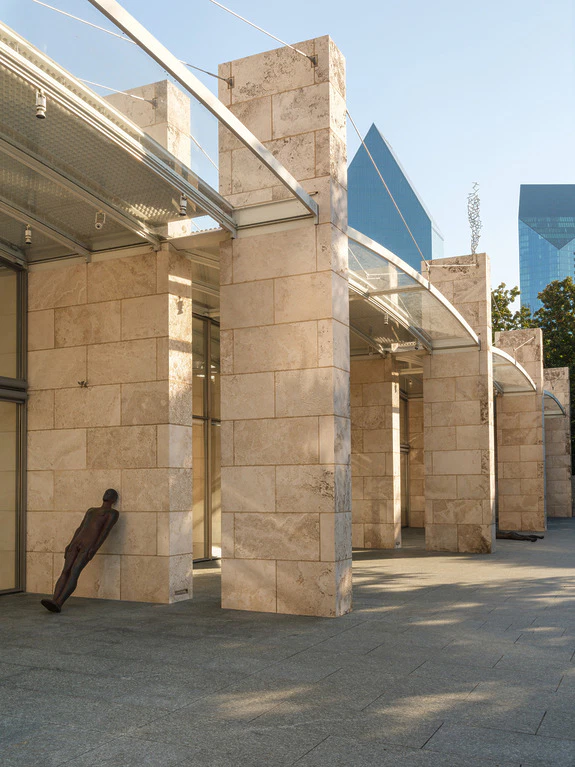 Modern architectural structure with stone columns and glass roof, featuring a leaning metal statue.