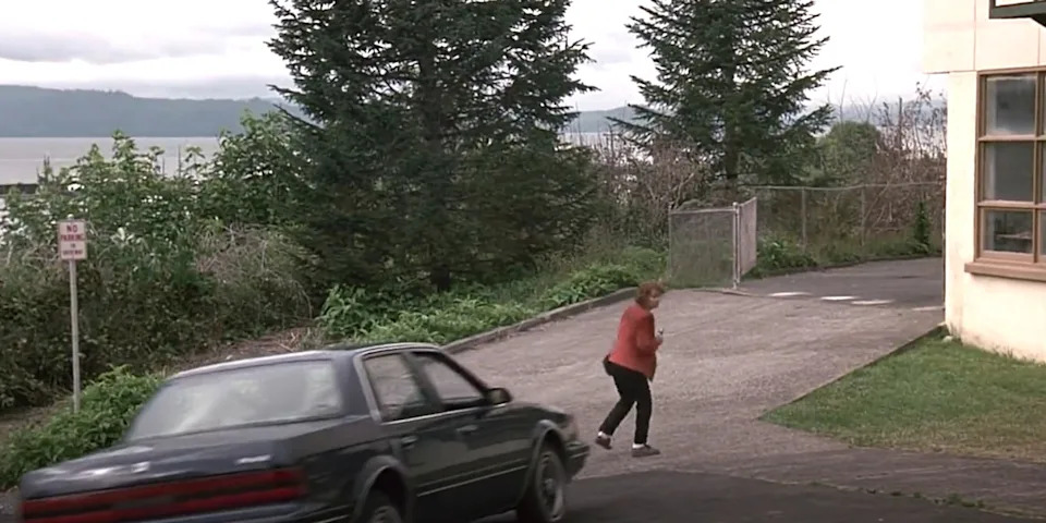 A person runs away from a parked car near a building, with trees and a body of water in the background