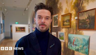 Oliver Jeffers is a man with black hair and beard who is standing in an art gallery in front of a wall of artworks. He is wearing a navy turtleneck top and coat.