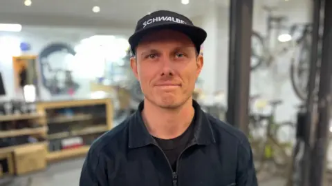 Max Geddes standing outside his bike shop. He is wearing a black cap and a black T-shirt with a dark jacket on top of it