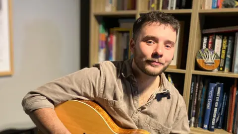 Andrew Sinclair/BBC Ross Stewart sitting down, holding a guitar, in front of a set of full bookshelves. He is looking directly at the camera and is smiling slightly. He has short dark brown hair and a neatly trimmed lighter brown beard. He is wearing a light brown casual shirt, open at the neck. Only the top of the guitar is visible, his right arm is over the top of the instrument.