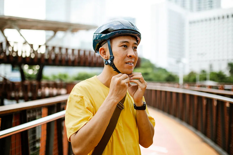 A person fastens a bike helmet while standing on a bridge in an urban area, surrounded by buildings in the background
