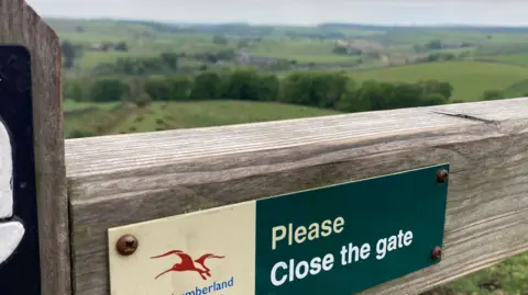 A cream and green Northumberland National Park: Please close the gate sign is nailed to a wooden gate. Lush, green fields stretch out behind it.