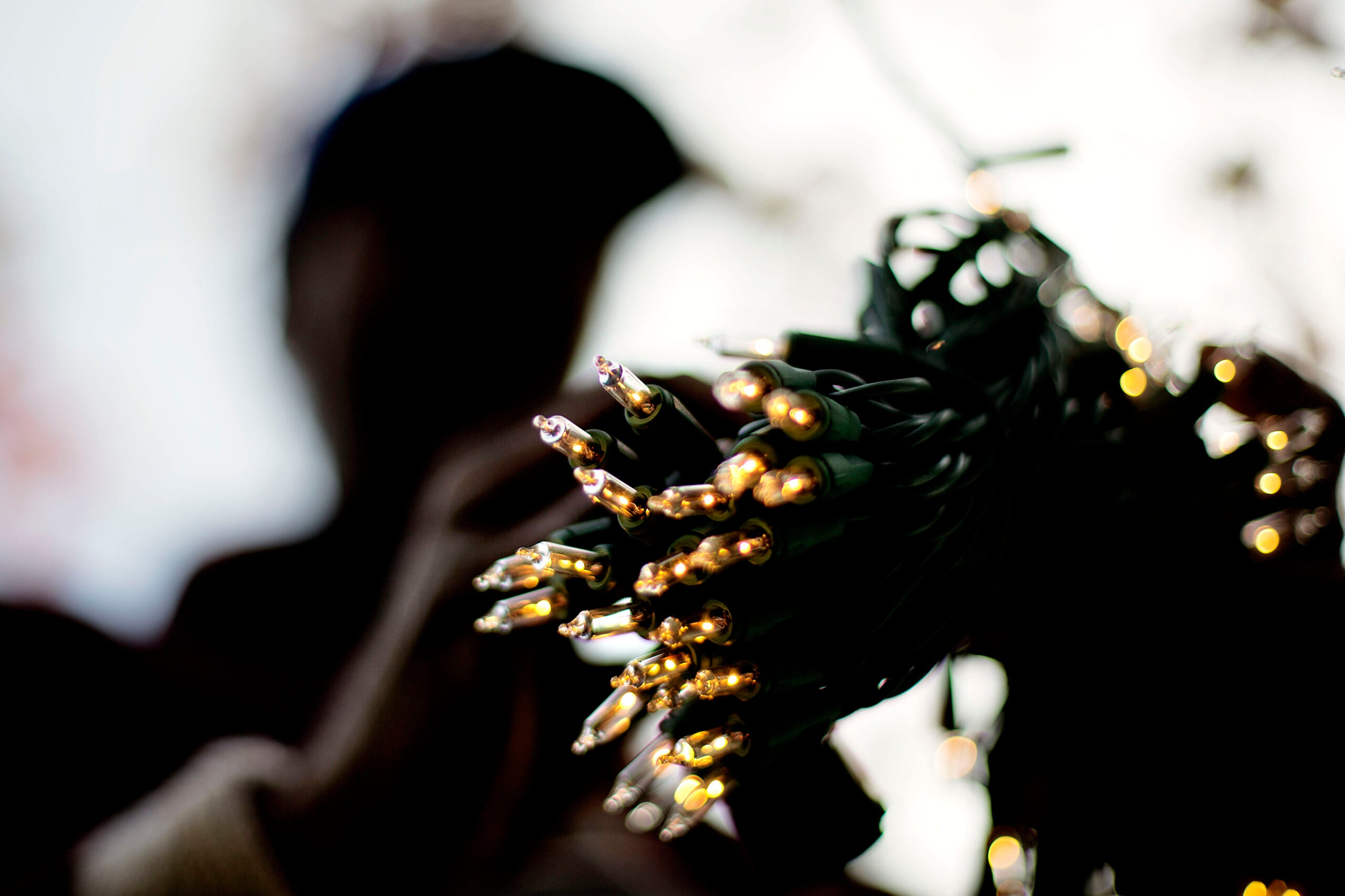 A bundle of lit string lights held in the foreground, with a blurry figure and light background behind.