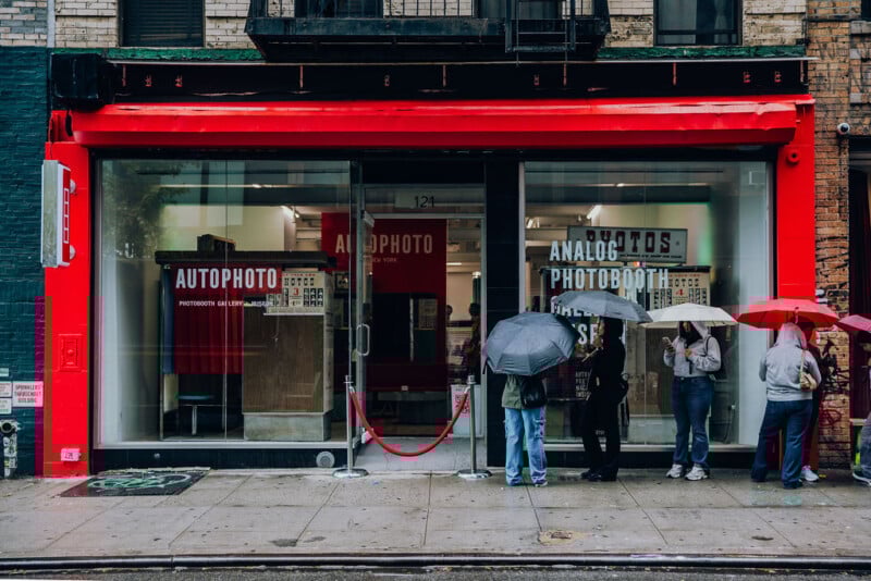 People with umbrellas stand outside a storefront with a bright red frame and glass windows. The shop displays signs reading “AUTOPHOTO” and “ANALOG PHOTOBOTH.” A red rope barrier stretches across the entrance.