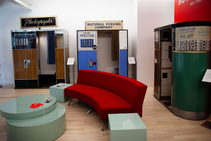 A modern room with vintage photo booths and vending machines against the wall, featuring a curved red couch and glass tables on a light wood floor.