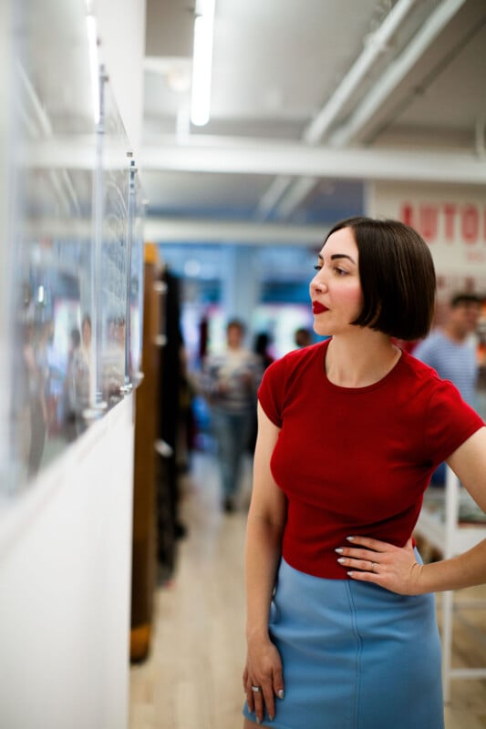 A woman with short dark hair, wearing a red top and light blue skirt, stands indoors, looking intently at artwork on a wall, with people and bright lights in the background.