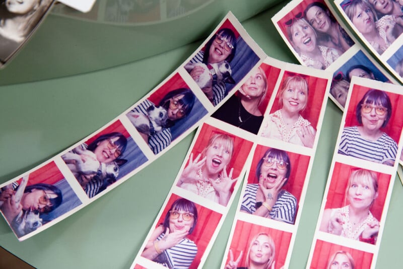 Three strips of photo booth pictures featuring women making playful faces and poses, some with a small dog. The strips are laid out on a green surface. The background of the photos is red.