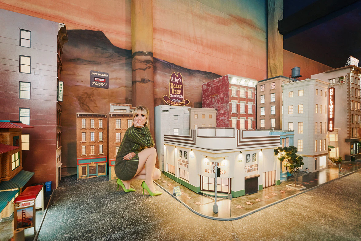 Woman in green dress and matching heels crouching beside a detailed miniature city display with buildings and street decorations.