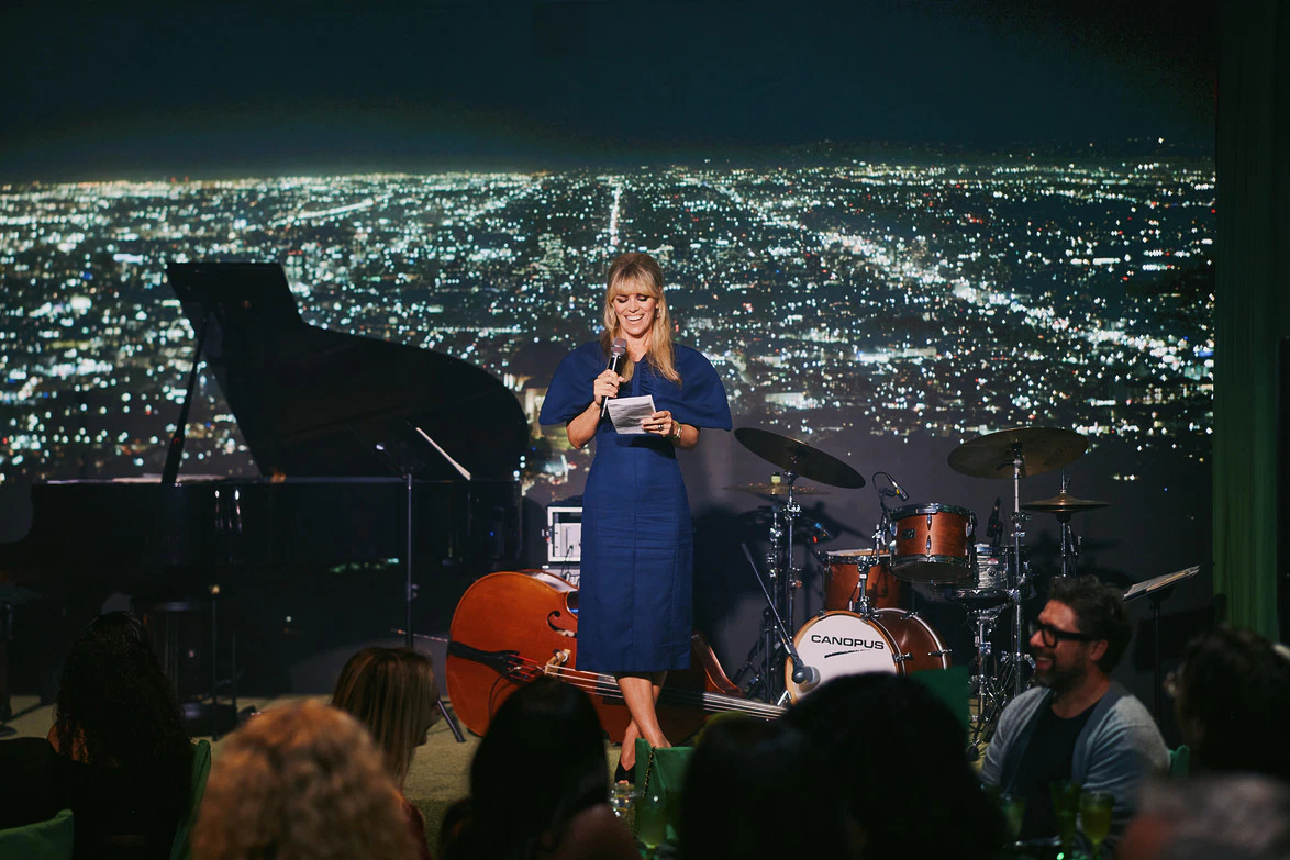 Comedian performs stand-up on stage with city skyline backdrop, microphones in hand and audience in foreground.