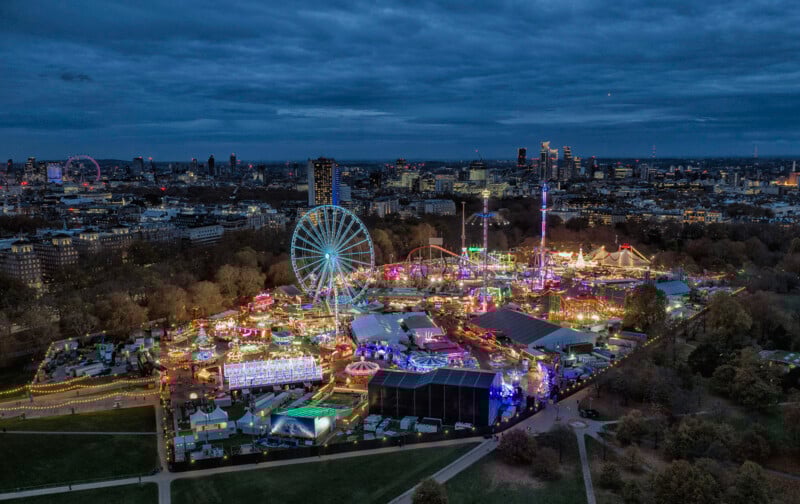 Aerial view of a brightly lit fairground at night, featuring a large Ferris wheel and various amusement rides, surrounded by city buildings under a cloudy sky.