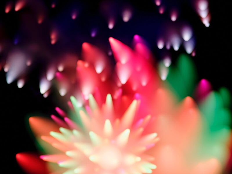 Abstract image with colorful, spiky shapes resembling bursts of light or fireworks in shades of red, green, pink, and white against a dark background. The lights are soft and blurred, creating a dreamy effect.