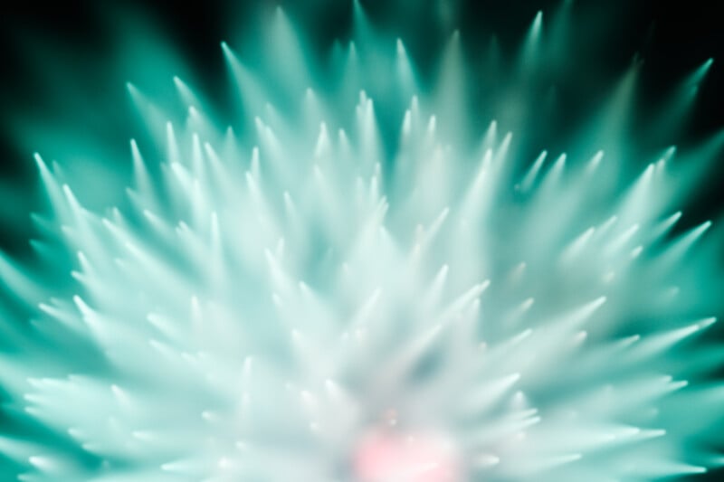 Abstract close-up image featuring soft, turquoise-white spikes radiating outward, creating a dreamy, glowing effect with a hint of pink at the center against a dark background.