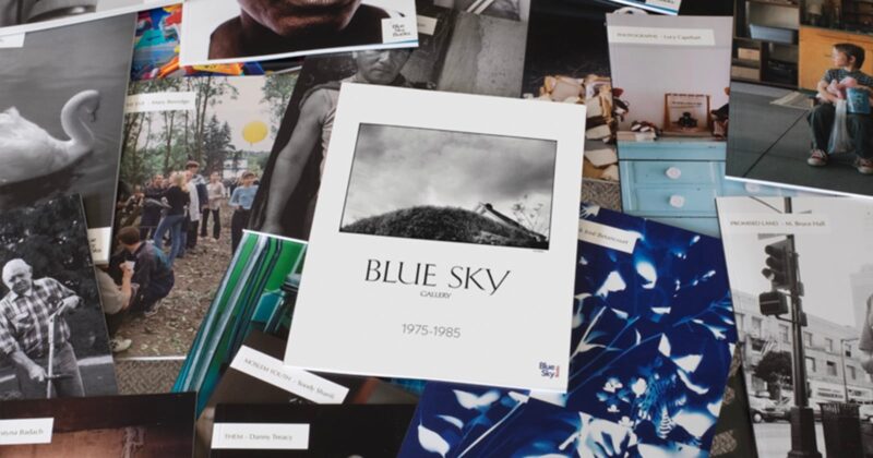 A variety of photography exhibition flyers and postcards are spread out, with a central catalog titled "Blue Sky Gallery 1975-1985" featuring a black-and-white photo of a hill with a house at the top.