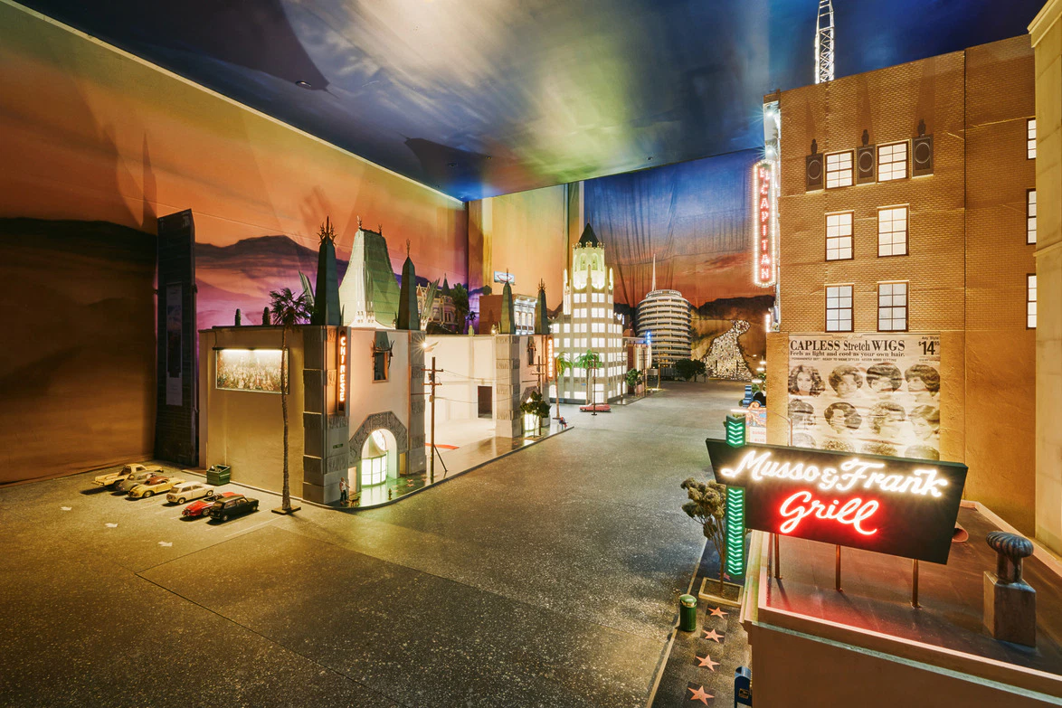 Model of a city street with buildings, neon signs, and vintage cars under a colorful sky backdrop at night.