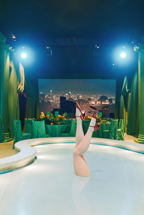 Person upside down in a pool with legs in red heels, indoor setting with cityscape background, green curtains and lights.