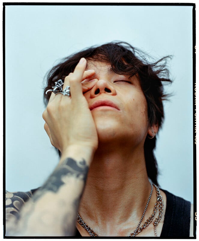 A person with tousled hair closes their eyes and covers part of their face with a tattooed hand, wearing silver rings and layered necklaces, against a plain light background.