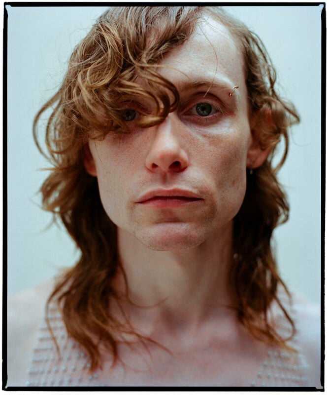 A person with fair skin and wavy, shoulder-length brown hair looks directly at the camera. One eye is partly covered by hair. They wear a faint expression and a sleeveless, textured top. The background is neutral and plain.